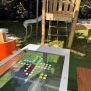 HPL Bench Chess and ludo game cu piese colorate pe o masă de joc în aer liber lângă echipament de joacă din lemn.